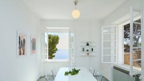 A bright, white dining room with a table and chairs, a view of the sea through an open window, and a ceiling light. - Hedera Estate, Hedera A30 (Dubrovnik)