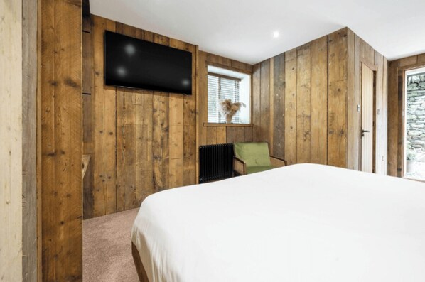 A bedroom with a wooden wall paneling, a bed, a green chair, a window with blinds, and a flat-screen TV mounted on the wall. - The Retreat, Windermere (windermere)