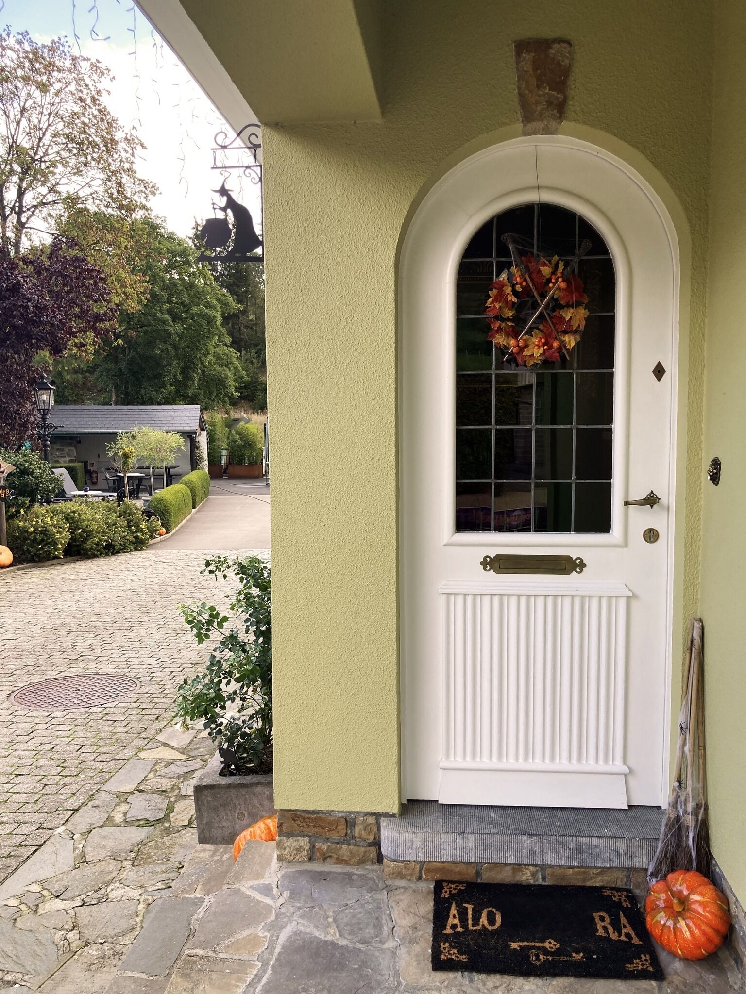 A white door with a glass panel and a wreath, a stone-paved entrance, a doormat, and a broom leaning against the wall.