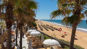 En terrasse ved stranden med udendørs siddepladser og parasoller, med udsigt over en sandstrand og havet.