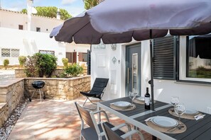 An outdoor patio with a table set for dining, a grill, and a black umbrella providing shade.