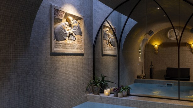 Un moderno espacio de spa con una amplia piscina interior, paredes con azulejos y dos esculturas iluminadas en la pared.