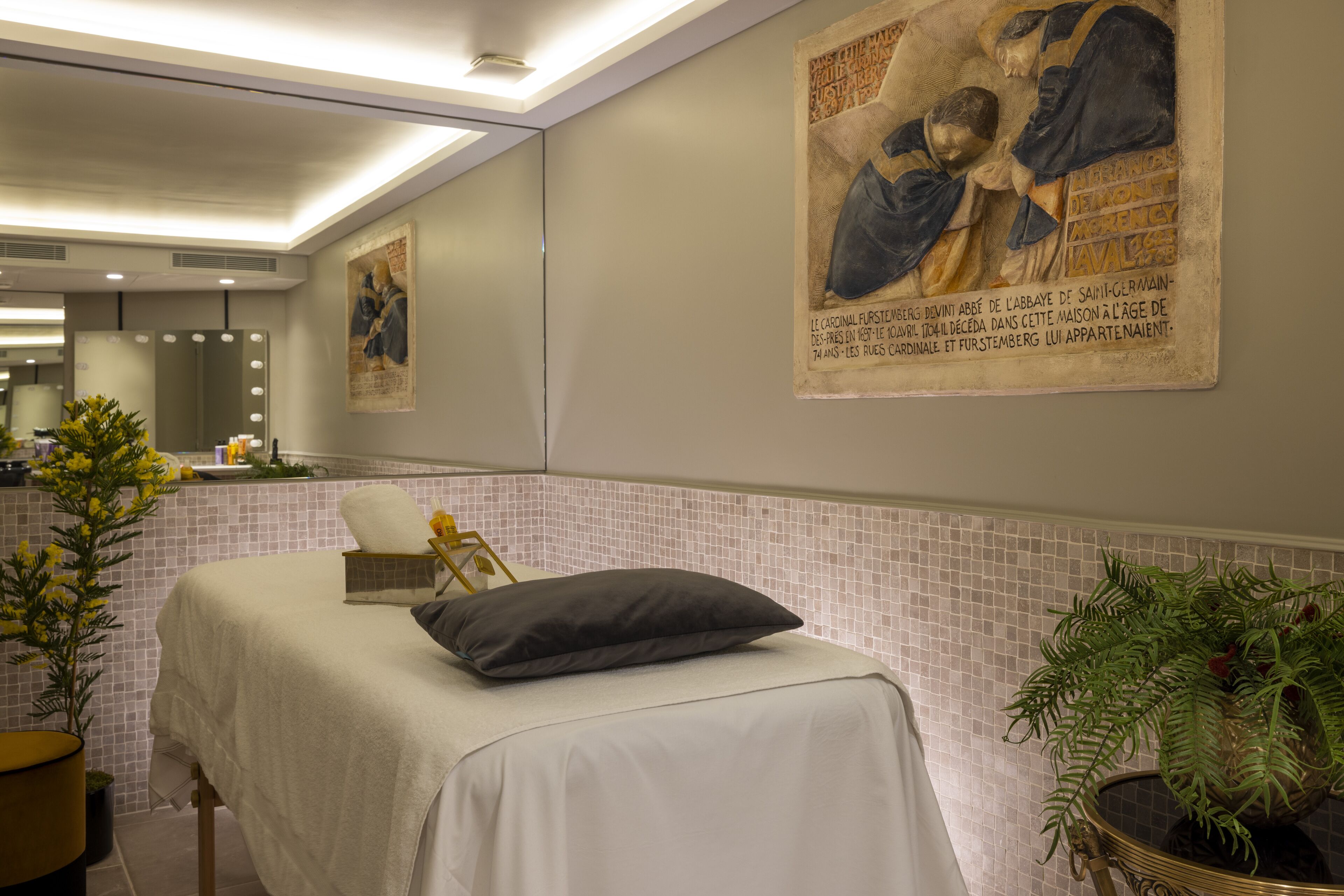 A room with a bed, a mirror, a potted plant, and a wall with a historical painting.
