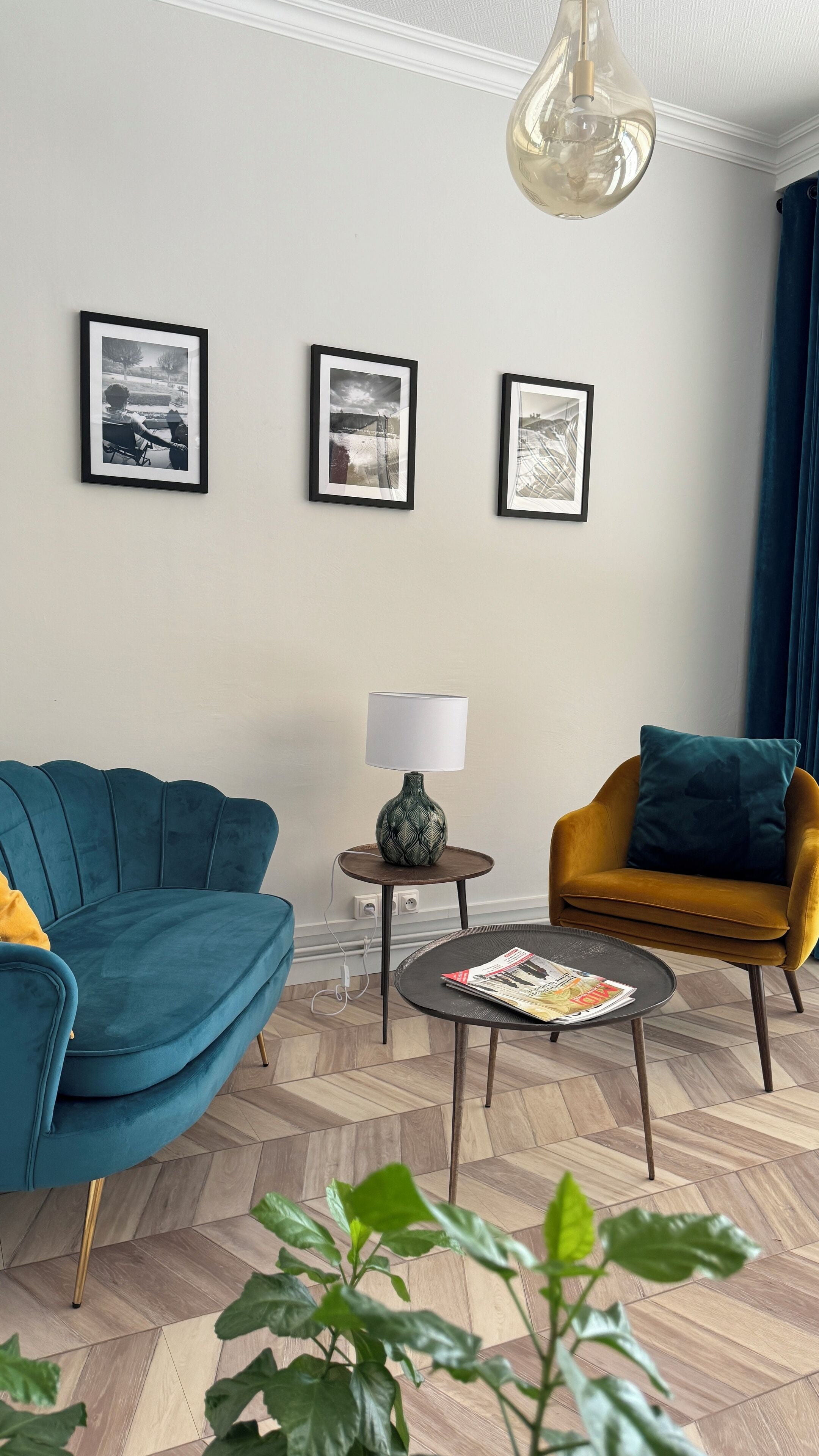 A living room with a teal sofa, a mustard armchair, a small round table with a lamp, and three framed pictures on the wall.