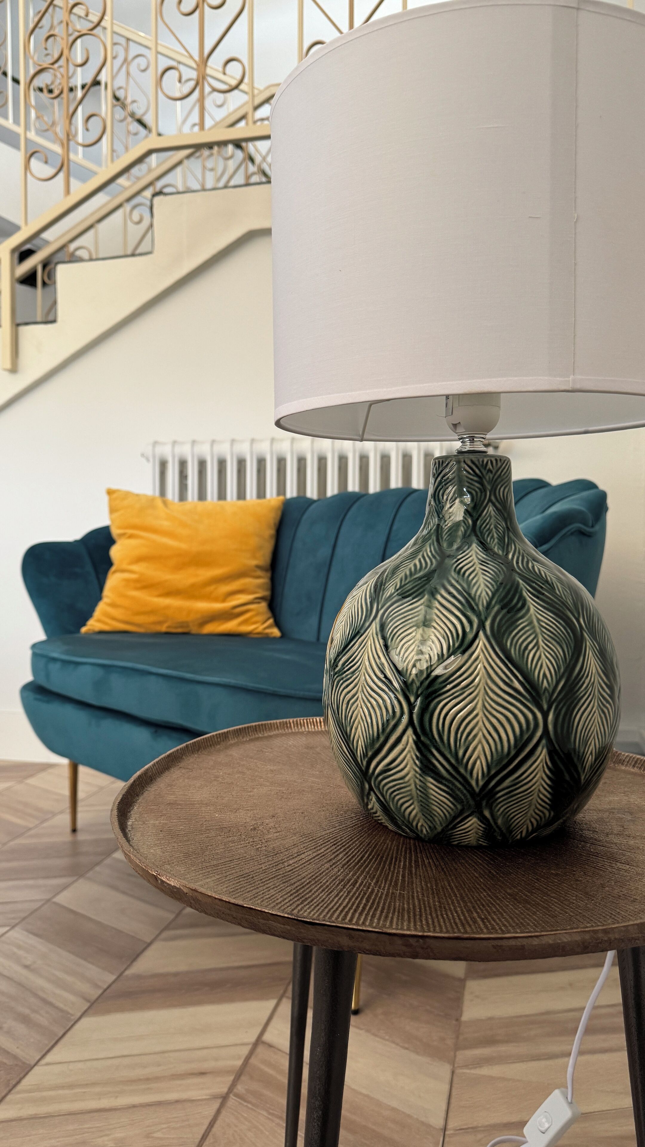 A modern living room with a decorative lamp on a round table, a teal sofa with a yellow pillow, and a staircase with a metal railing in the background.