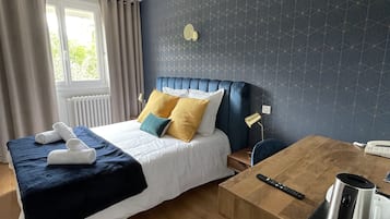 A hotel room with a bed, a wooden table with a kettle, a remote control, and a window with curtains.