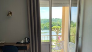 A hotel room with a bed, a desk, a chair, and a view of a balcony with a tree and a lake. - Hôtel Restaurant du Lac (Boussens)