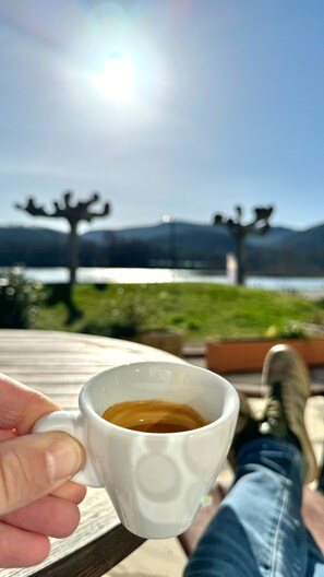 A hand holding a white mug with a beverage, overlooking a scenic view of a lake and mountains. - Hôtel Restaurant du Lac (Boussens)