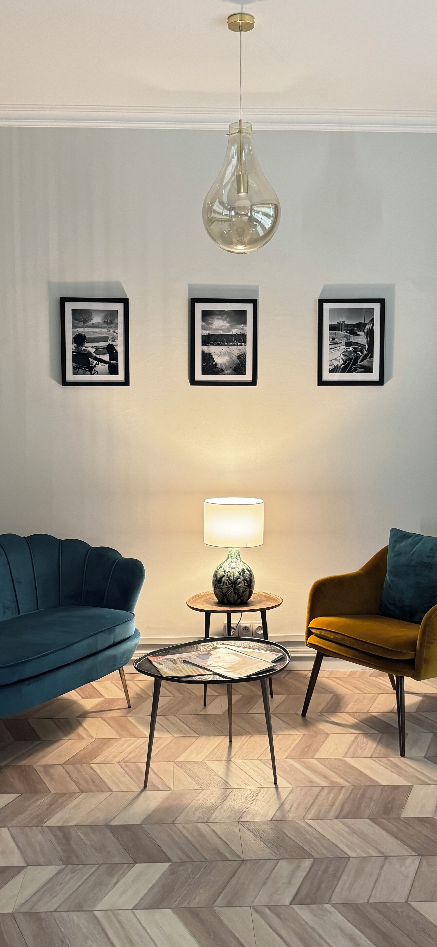 Uma sala de estar moderna com um sofá azul, uma poltrona mostarda, uma mesa de centro redonda e um lustre. Três fotografias emolduradas estão expostas na parede.