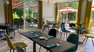 Un comedor con mesas y sillas, ventanas amplias con cortinas amarillas y mobiliario exterior visible a través de las puertas de vidrio.