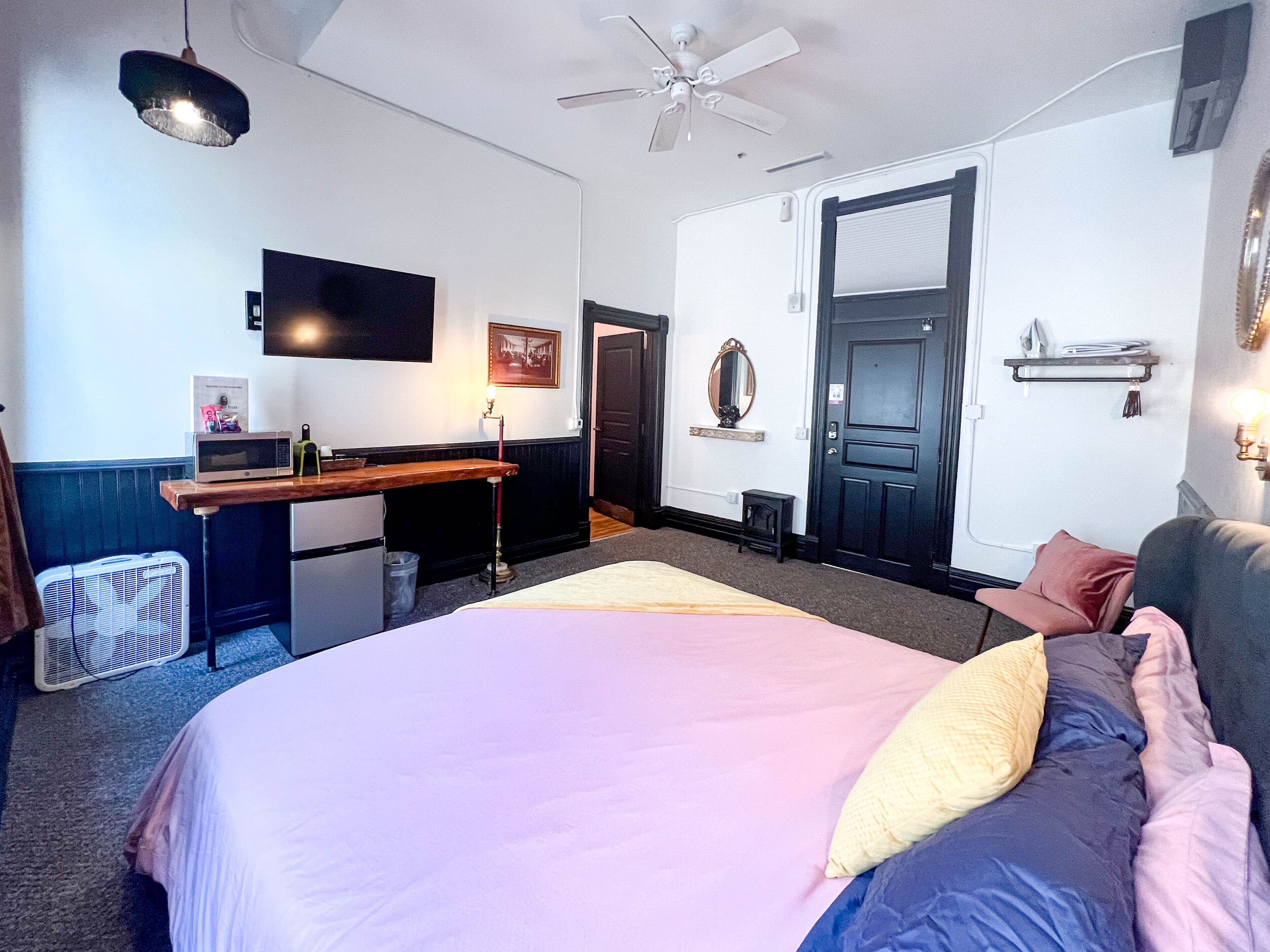 A bedroom with a bed, a desk, a chair, a fan, and a door.