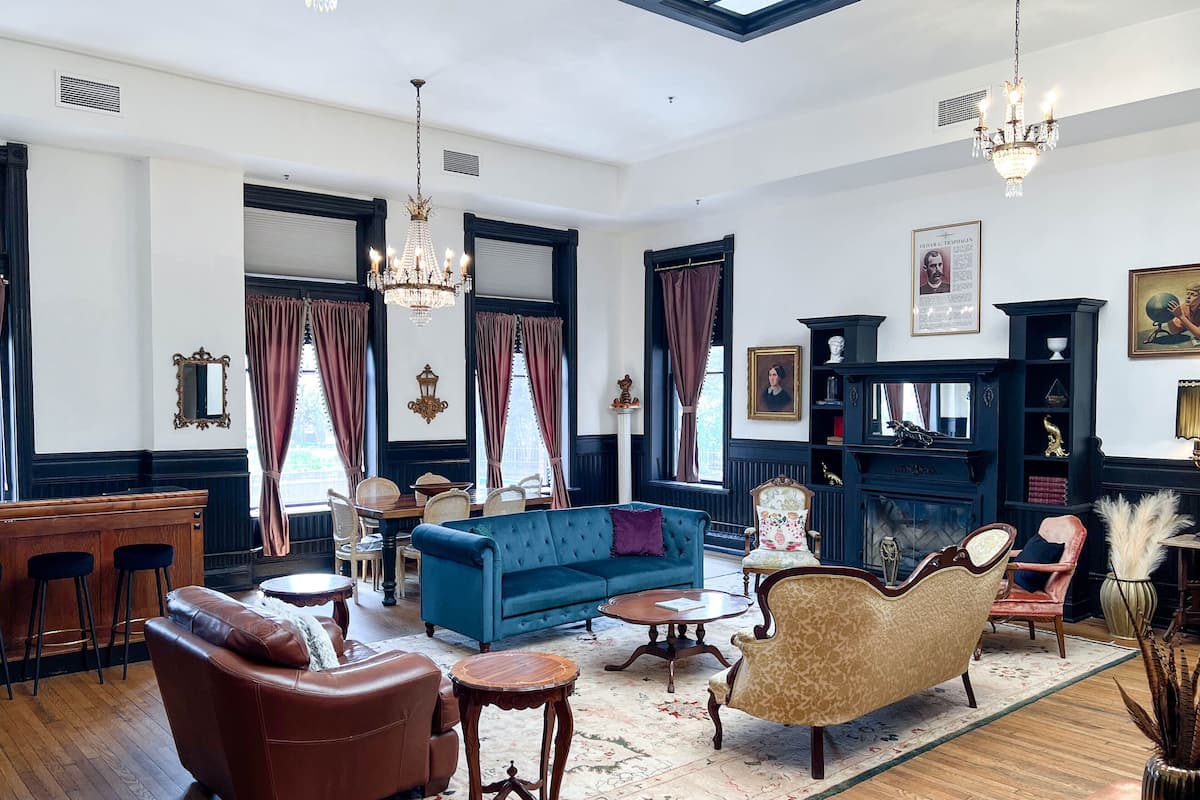 A spacious living room with a blue sofa, a brown leather couch, and a patterned rug. There are two chandeliers, a fireplace, and a bookshelf with various items.