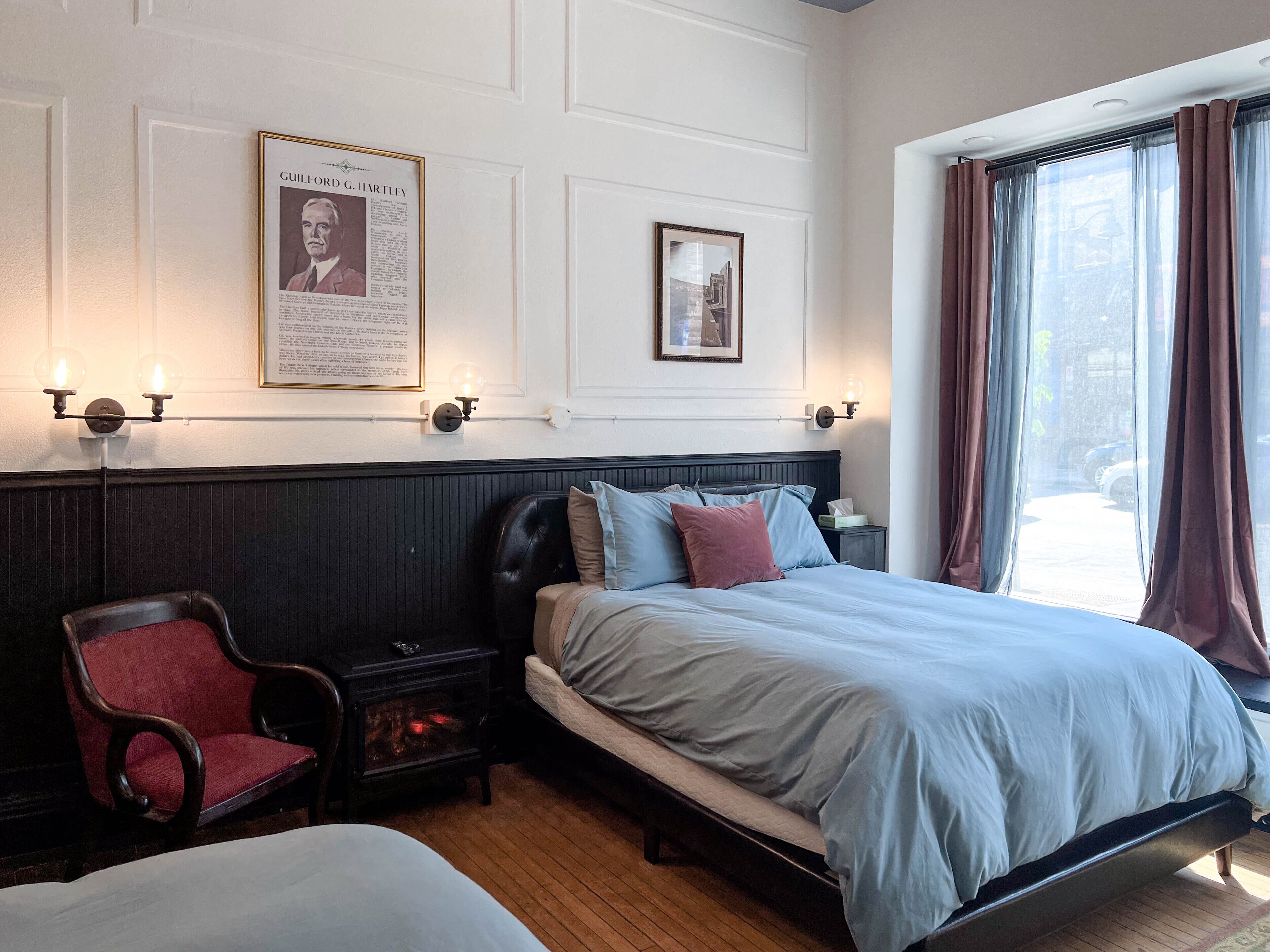 A hotel room with a bed, a chair, a nightstand, wall-mounted lamps, and framed pictures on the wall.