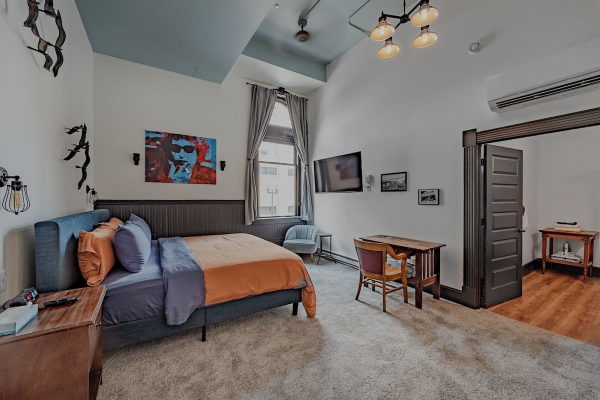 A bedroom with a bed, a desk, a chair, a TV, and artwork on the walls.