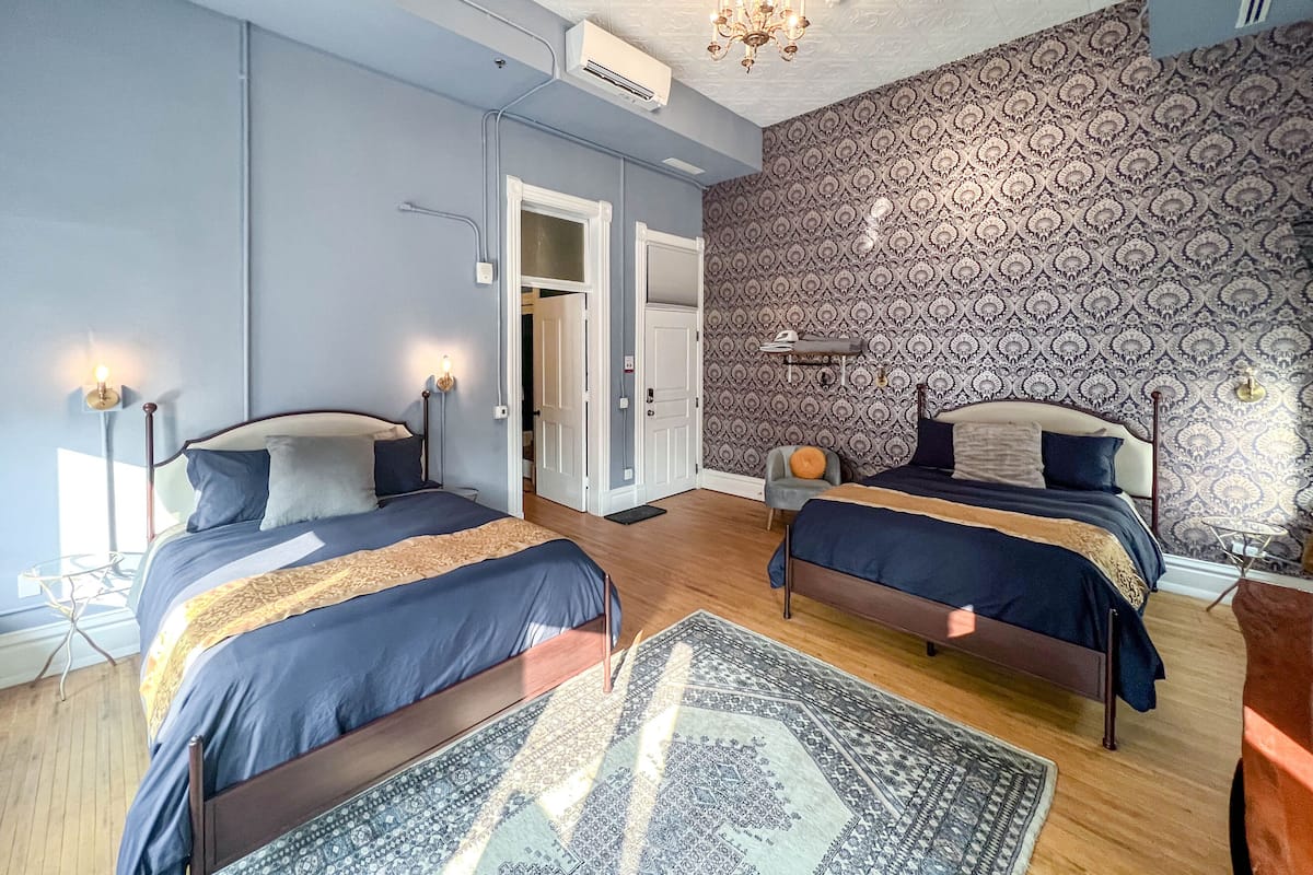 A modern bedroom with two beds, patterned wallpaper, a chandelier, and a visible door leading to another room.