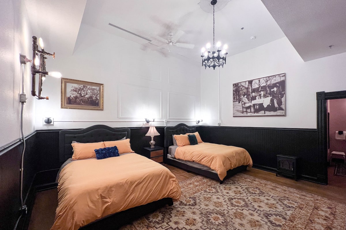 A hotel room with two beds, a chandelier, a patterned rug, and wall-mounted artwork.
