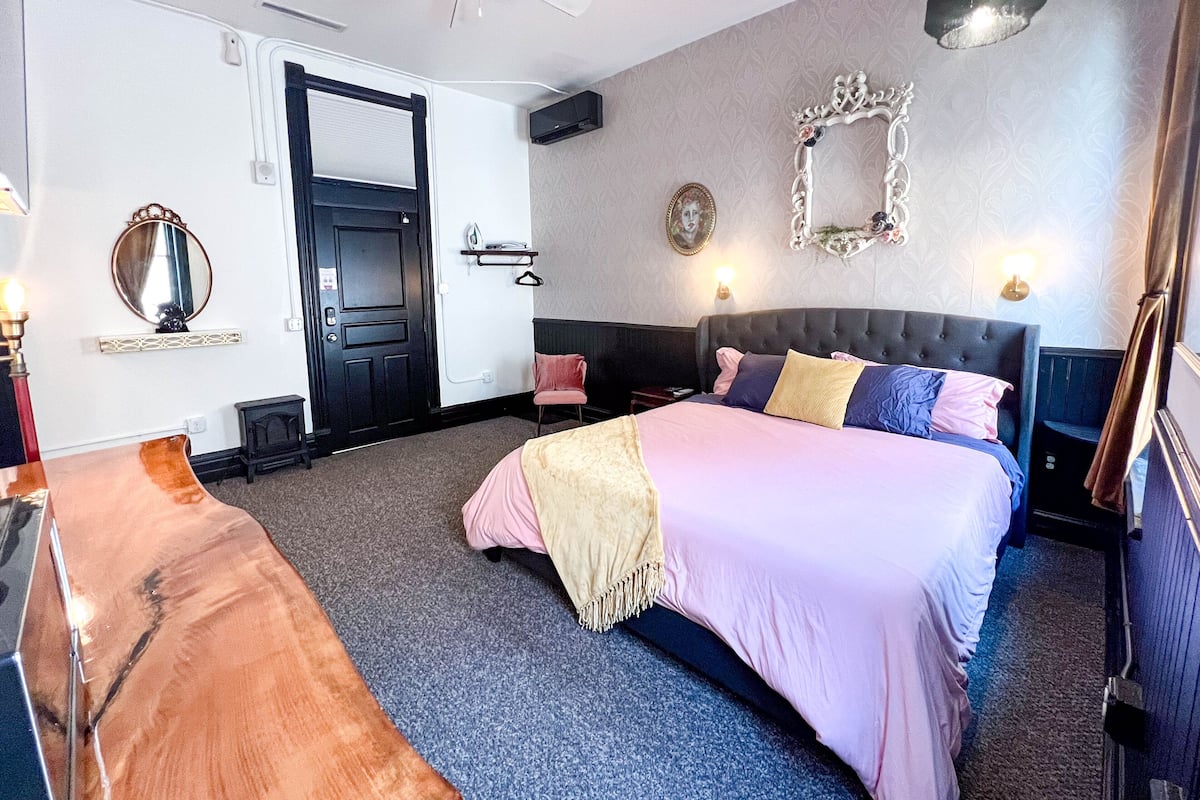 A bedroom with a large bed, a bench, a door, a mirror, and a wall-mounted light fixture.
