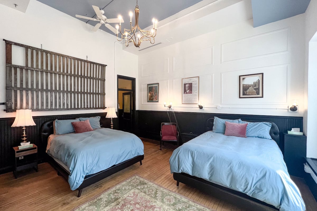 A hotel room with two beds, a chandelier, a wooden headboard, a patterned rug, and framed pictures on the wall.
