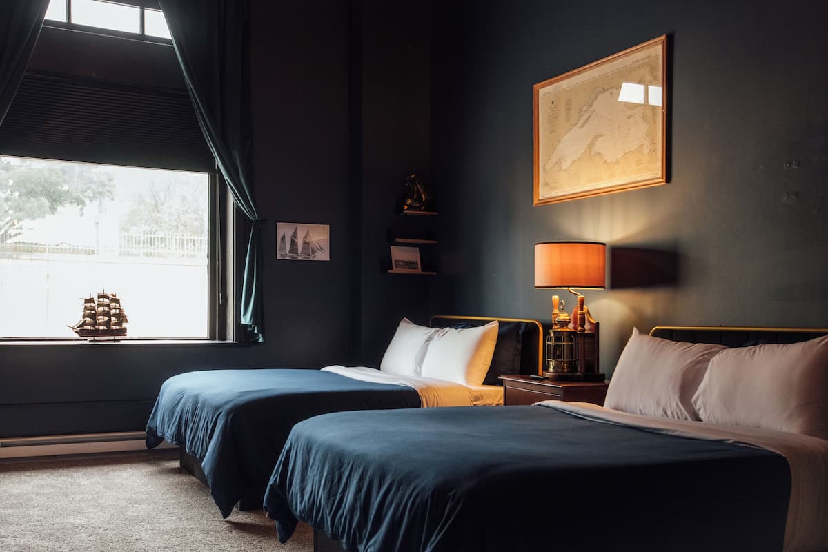 A hotel room with two beds, a large window with curtains, a map on the wall, and a lamp on the nightstand.