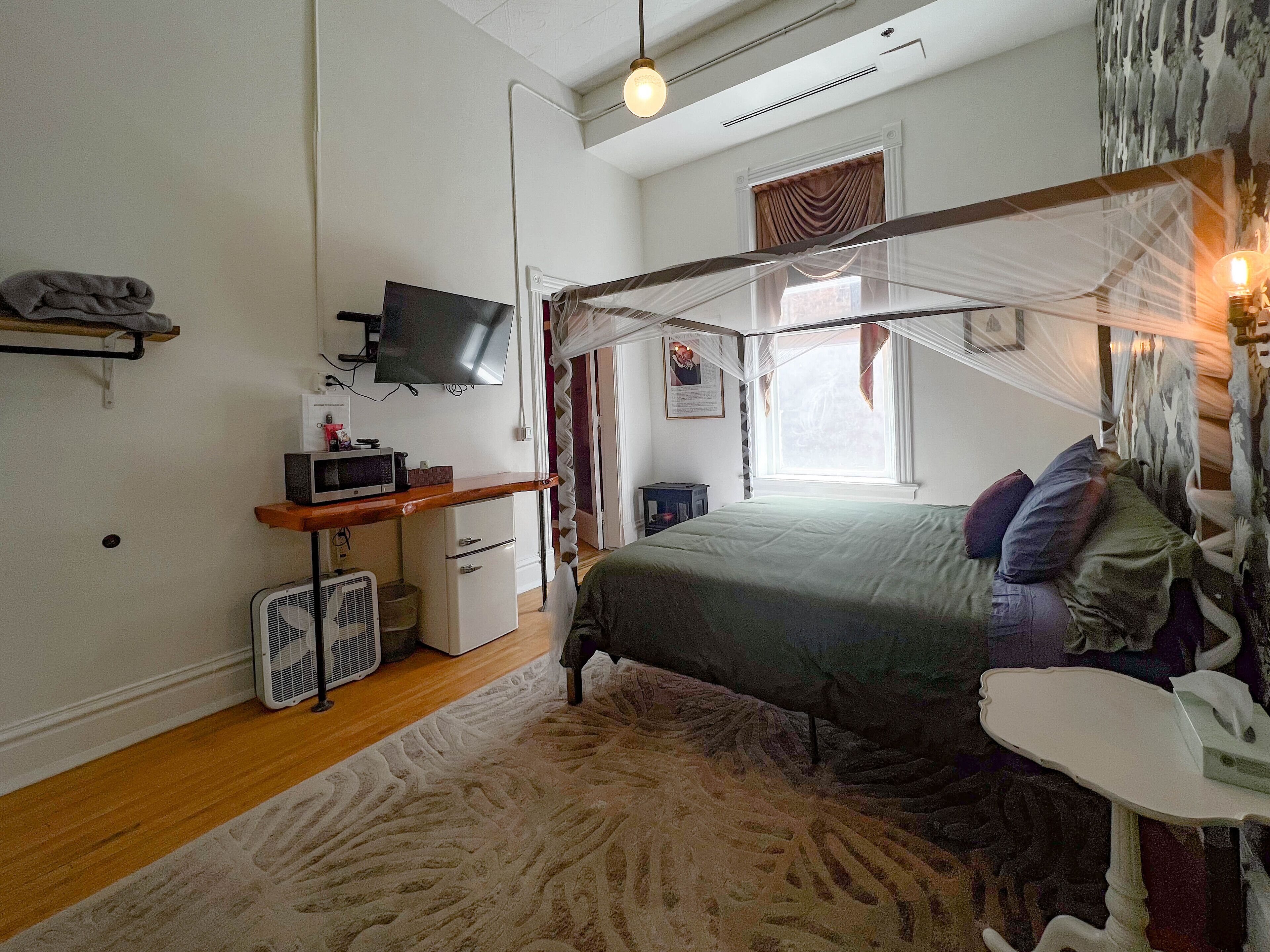 A bedroom with a bed, a desk, a television, a small heater, and a window with curtains.