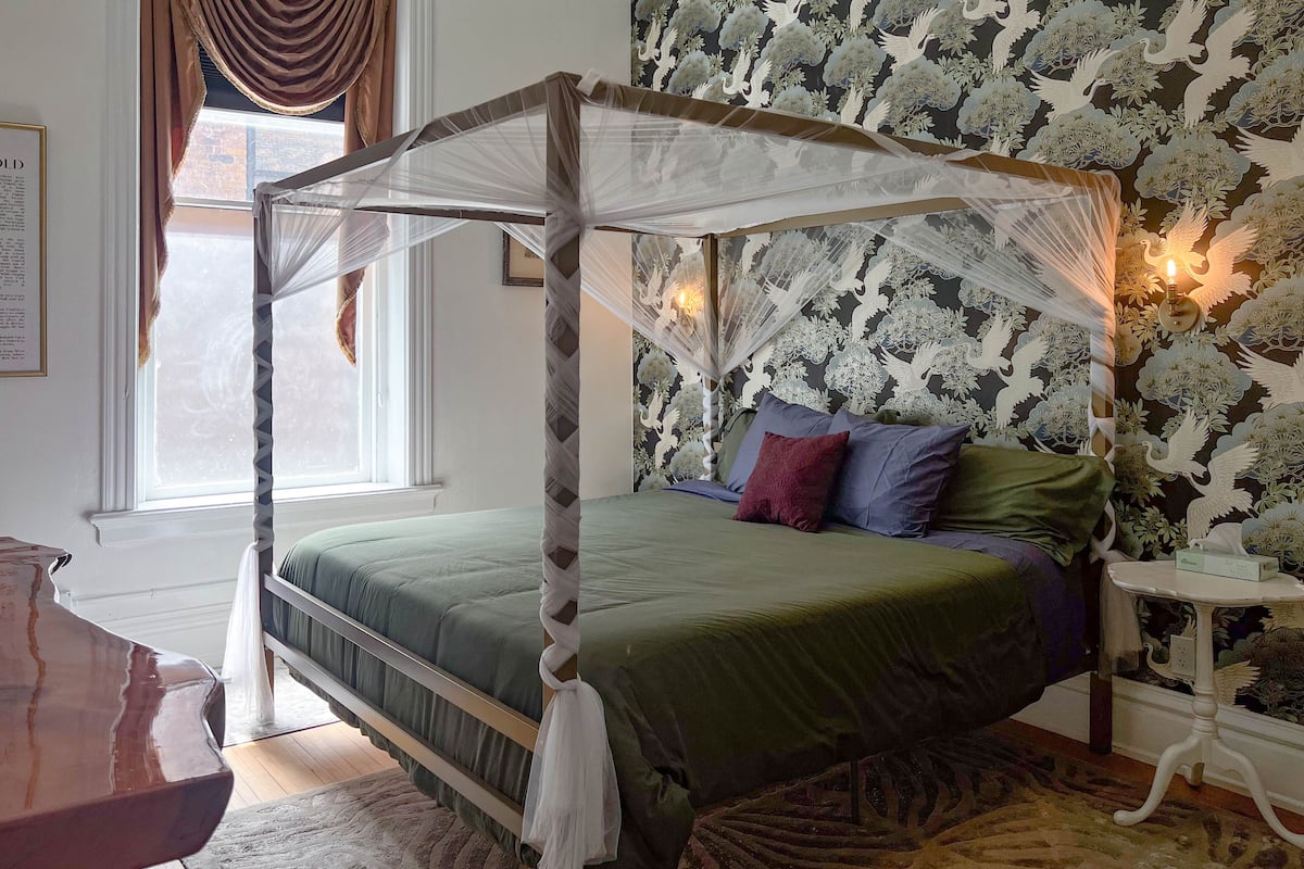 A four-poster canopy bed with a green bedspread and multiple pillows, a patterned rug, and a small table with a lamp.