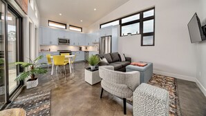 A modern kitchen with light blue cabinets, a stainless steel refrigerator, and a microwave. There is a dining area with yellow chairs and a table. The living room has a gray sofa and a patterned rug.