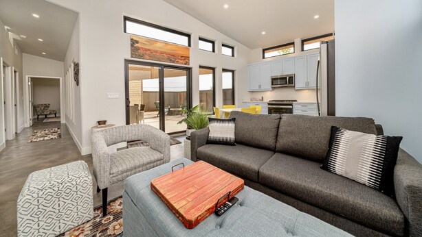 A modern living room with a grey sofa, a patterned ottoman, and a wooden coffee table. A kitchen with white cabinets and a microwave is visible in the background.