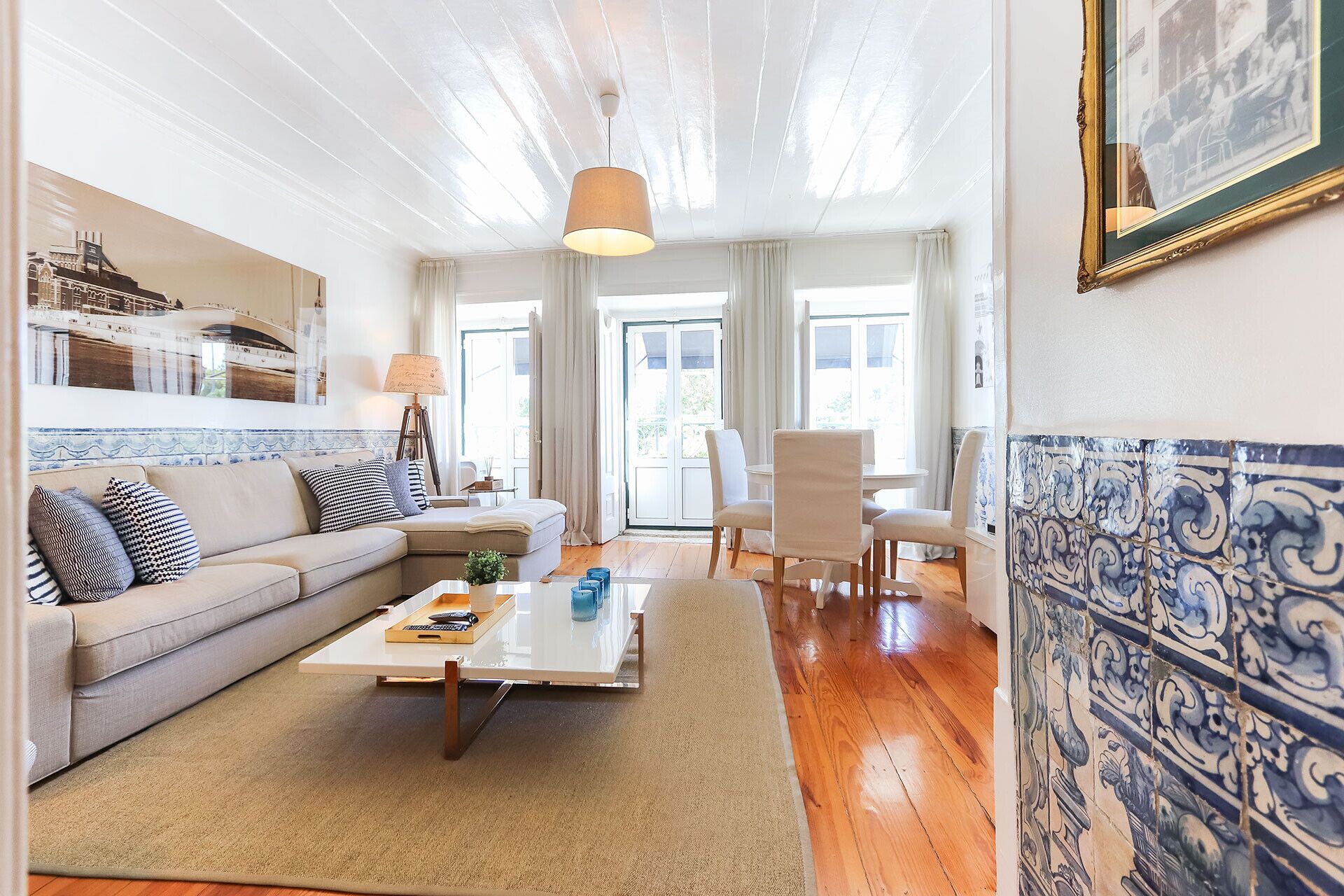 Una sala de estar con un sofá, una mesa de centro y sillas. Se ve una ventana con cortinas, una lámpara y una pared de azulejos.