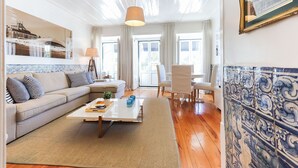A living room with a sofa, coffee table, and chairs. There is a window with curtains, a lamp, and a tiled wall. - Belem Prestige by Homing (Lisbon)