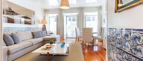Una sala de estar con un sofá, una mesa de centro y sillas. Se ve una ventana con cortinas, una lámpara y una pared de azulejos.