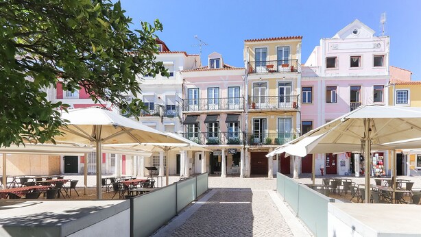 A cobblestone courtyard with outdoor seating under umbrellas, surrounded by pastel-colored buildings with balconies. - Belem Prestige by Homing (Lisbon)