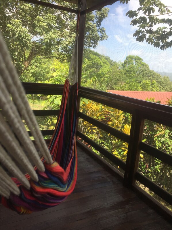 A hammock hanging on a balcony with a view of lush greenery and a red-roofed building. - Santa Cruz Cabins (San Ignacio)