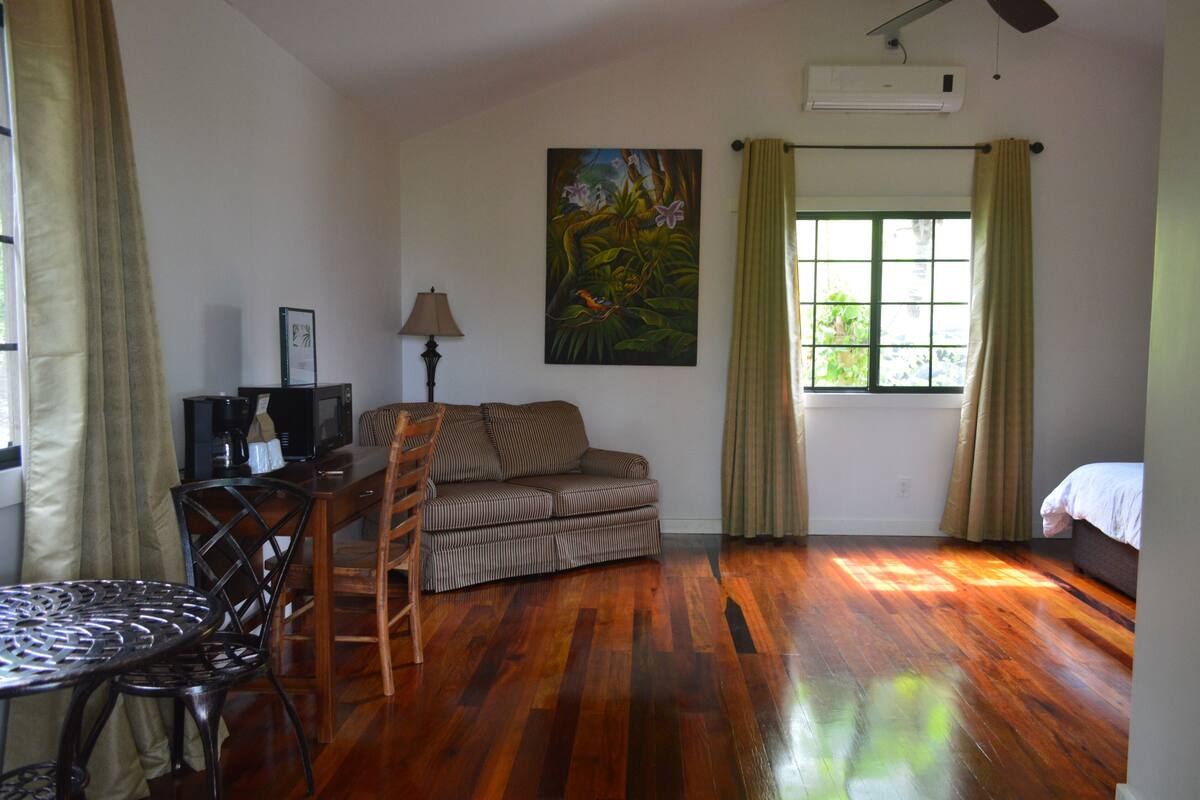 A room with a wooden floor, a sofa, a dining table with chairs, a painting on the wall, and a window with curtains.
