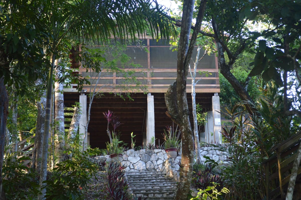 A traditional wooden structure with a stone staircase leading up to a covered entrance, surrounded by lush greenery.
