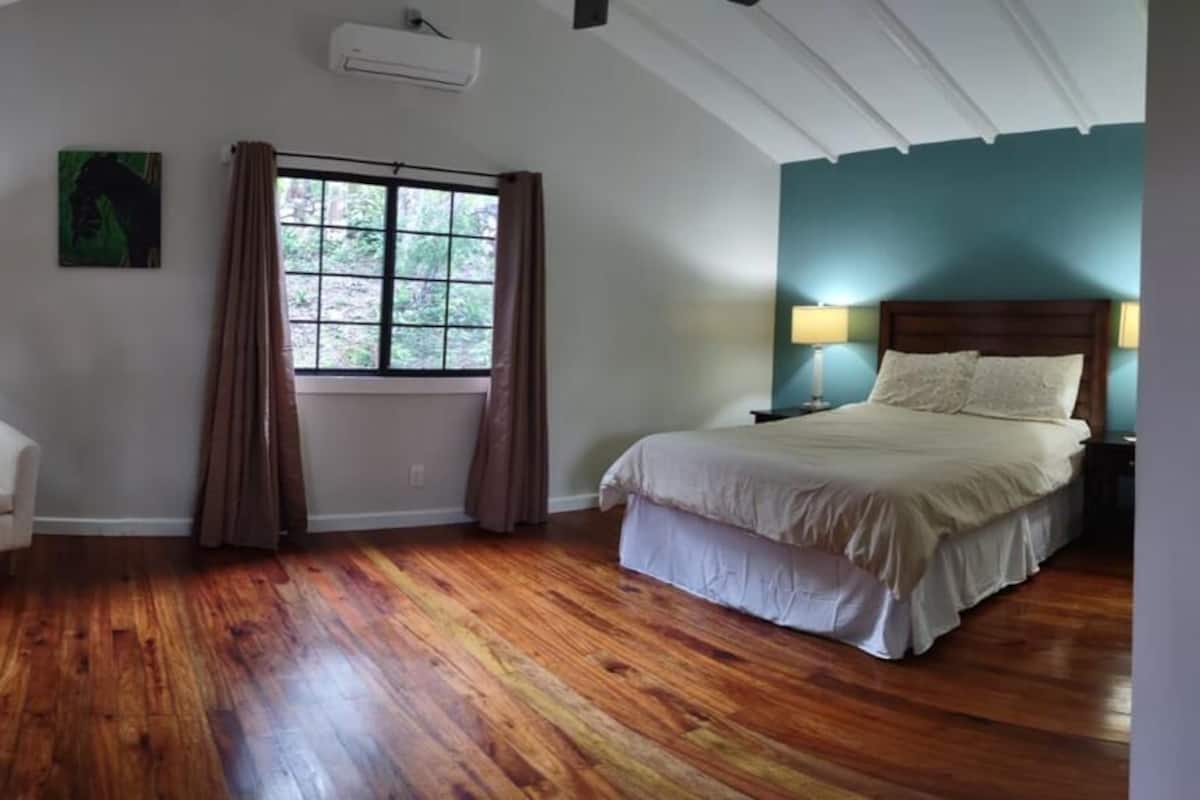 A bedroom with a wooden floor, a bed with white bedding, a chair, a window with curtains, and a wall-mounted air conditioner.