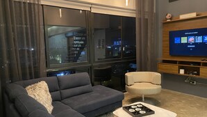 A modern living room with a dark grey sofa, a patterned armchair, and a coffee table with a game. A large TV is mounted on the wall, and there is a window with a view of the city at night.