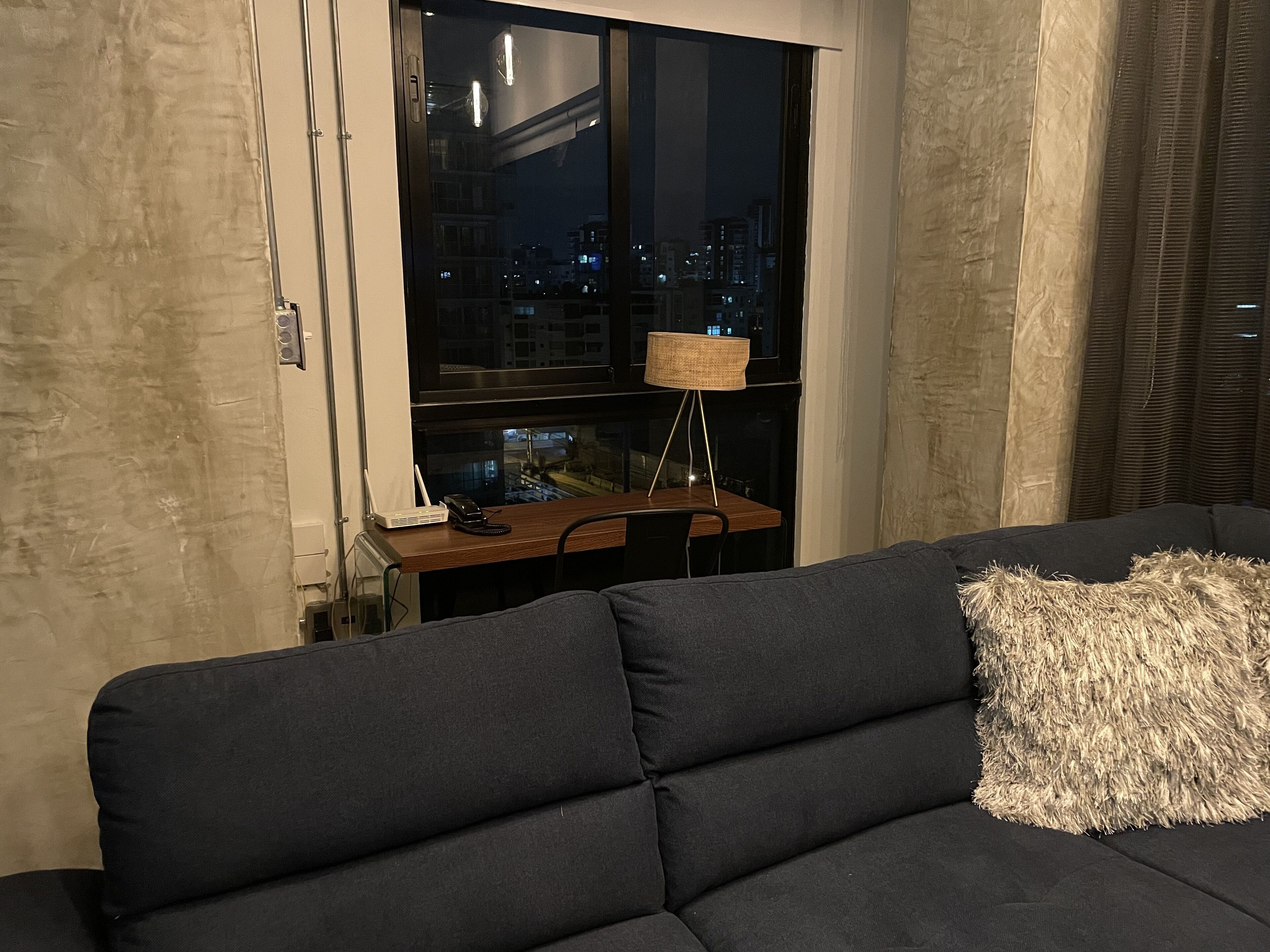 A modern living room with a dark grey sofa, a small wooden table, and a lamp. A window reveals a cityscape at night.