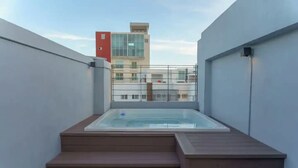 A rooftop terrace with a hot tub, wooden steps, and a view of surrounding buildings.