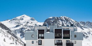 En modern byggnad med en symmetrisk fasad, stora fönster och ett platt tak, som kontrasterar mot en bakgrund av snötäckta berg och en klarblå himmel.