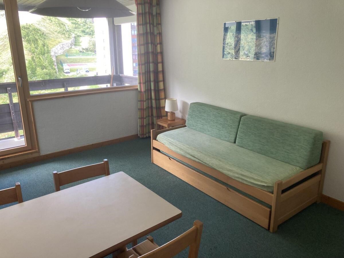 A room with a wooden table and chairs, a green sofa, a small bedside table with a lamp, and a window with curtains.