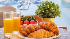 Ett frukostbord med croissanter, juice, jordgubbar och en krukväxt, med utsikt över en småbåtshamn och stadens silhuett.