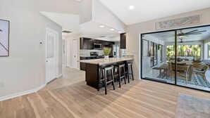A modern kitchen with a central island, bar stools, and a sliding glass door leading to a dining area with outdoor seating. - Waterfront Paradise with HTD Pool, Kayaks & Games (Cape Coral)