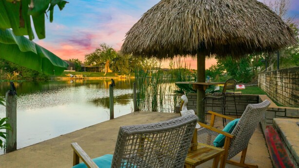 A tranquil outdoor seating area with a thatched umbrella, overlooking a calm body of water and a lush garden. - Waterfront Paradise with HTD Pool, Kayaks & Games (Cape Coral)