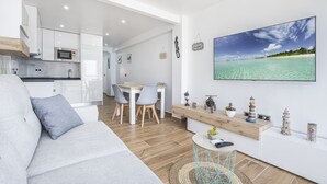 Ett modernt vardagsrum med en grå soffa, en matplats med ett träbord och stolar, ett kök med vita skåp och en mikrovågsugn samt en stor tavla med strandmotiv på väggen.