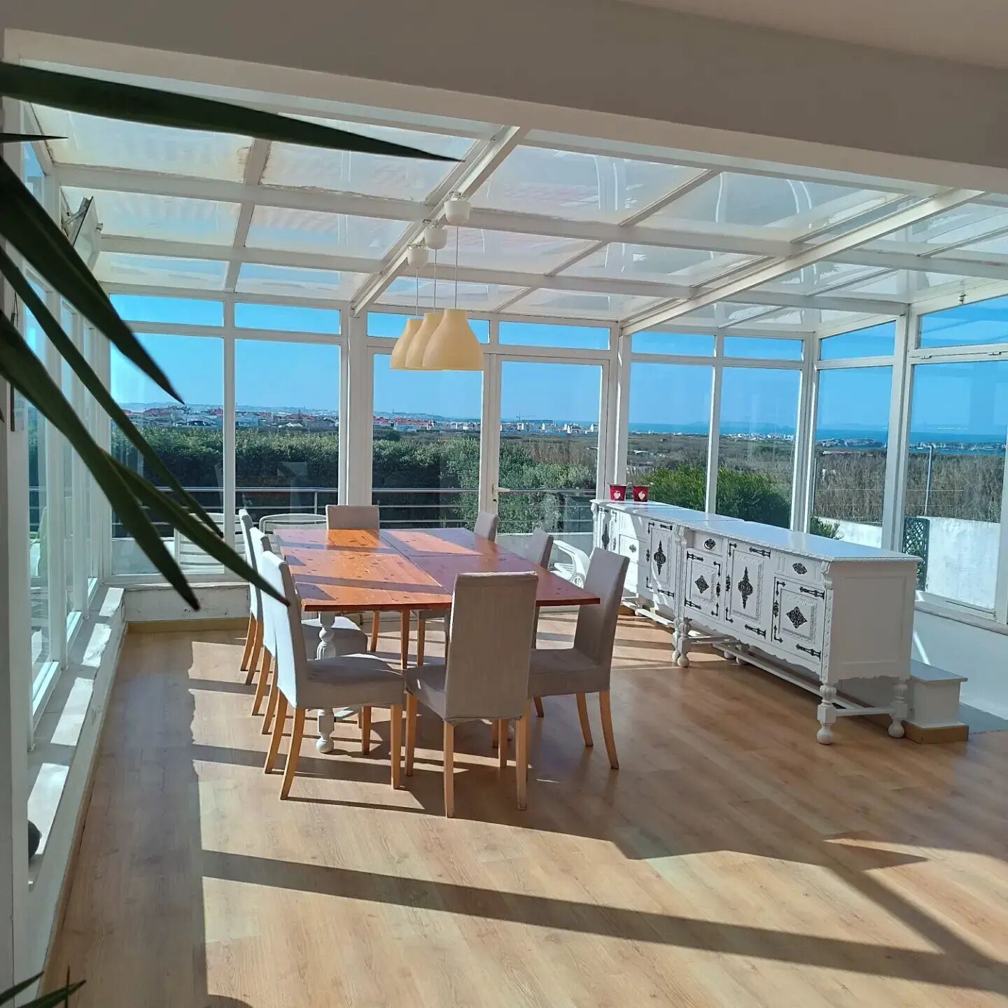 A sunlit room with a large table and chairs, a white cabinet, and a view of the landscape through large windows.