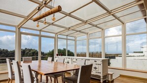 A spacious conservatory with a wooden dining table and chairs, large windows, and a view of the sea. - FreeSurfCamp (Peniche)