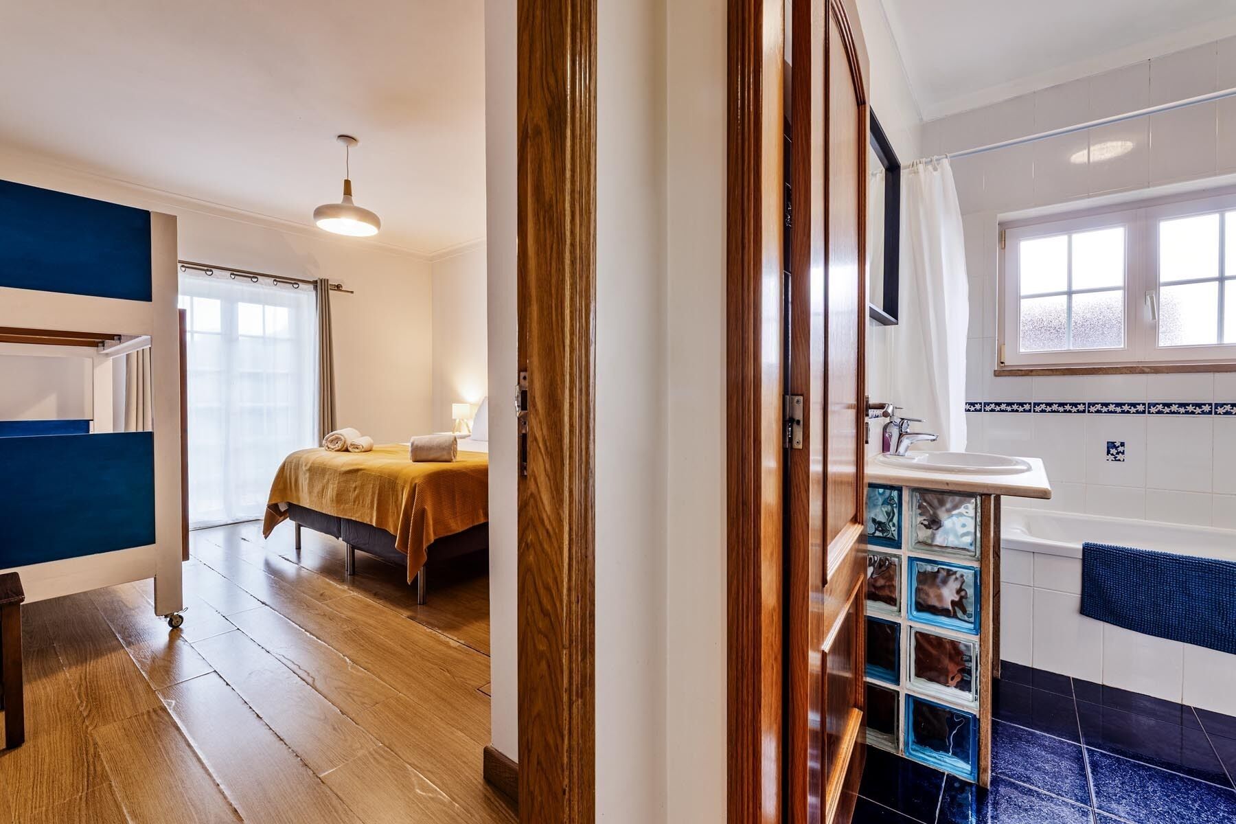 Una habitación de hotel con cama, mesita de noche, lámpara, ventana y baño con lavamanos y espejo.