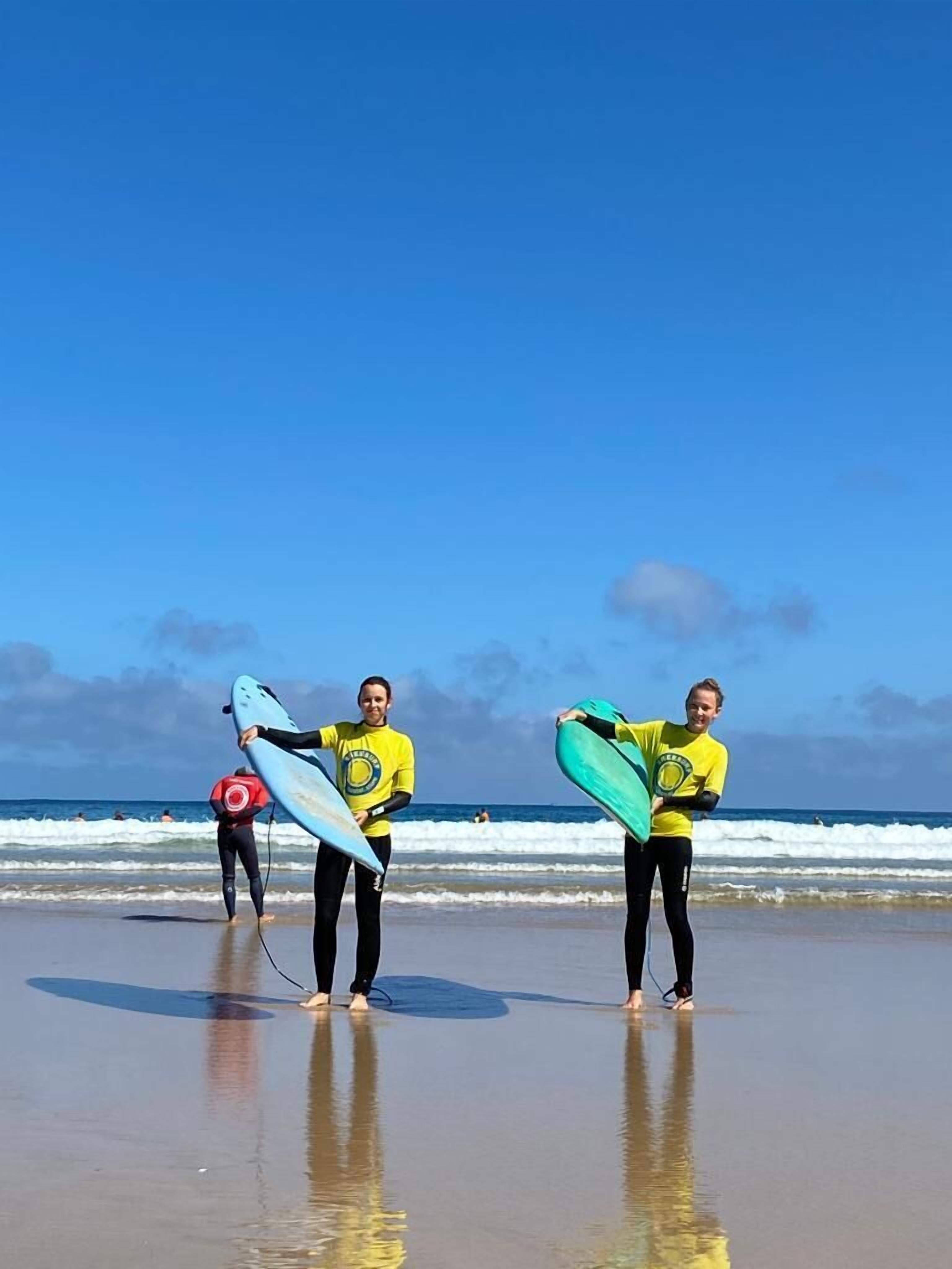 Zwei Personen am Strand mit Surfbrettern, im Hintergrund Wellen.