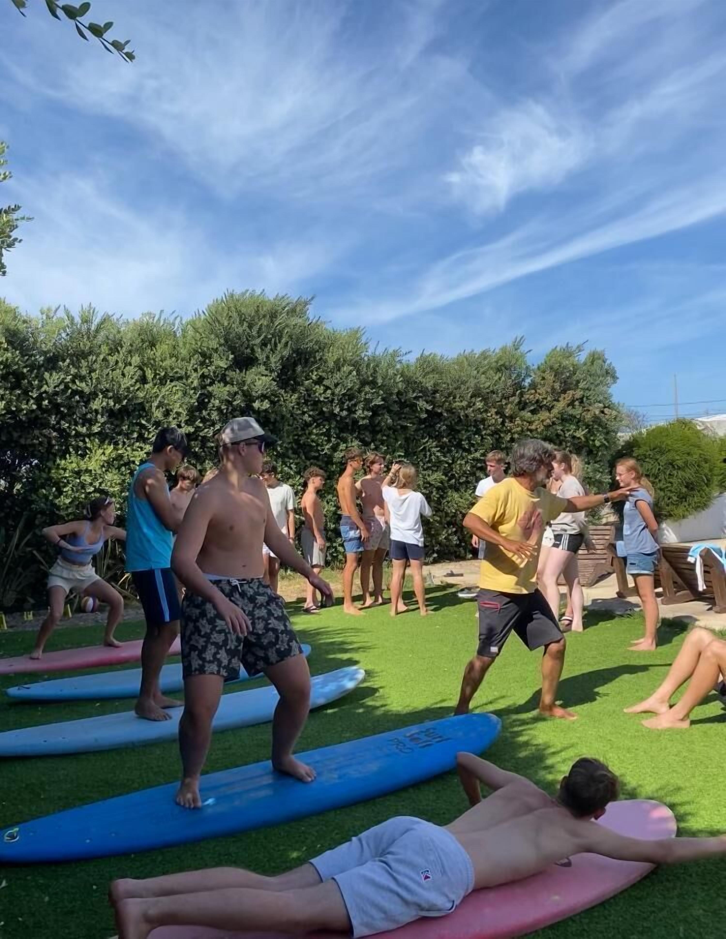 Un grupo de personas sobre tablas de surf en una zona cubierta de hierba, con árboles al fondo.