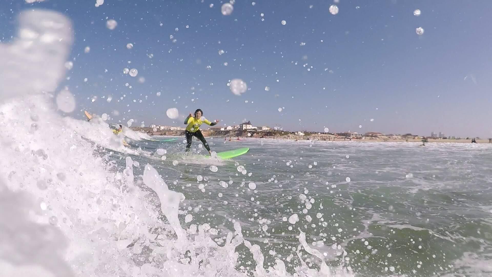 A person surfing on a wave with a clear view of the coastline and buildings in the background.